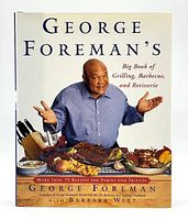 Front cover of George Foreman cookbook showing the author with grilled foods and kitchen utensils on table.