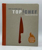 Front cover of the hardcover cookbook 'How to Cook Like a Top Chef' with a beige cover featuring a chef's knife cutout and text indicating recipes, techniques, and interviews from Bravo's hit show.