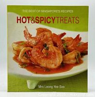 Front cover of the softcover cookbook featuring a plate of shrimp in spicy sauce on a green background with title text.