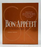 Front cover of The Bon Appetit Cookbook showing the orange hardcover with white and faint large letters spelling 'BOOK'.