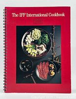 Front cover of the red softcover spiral-bound cookbook showing plated international foods.