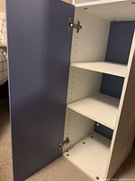 Open view of the shelving unit showing blue door with metal hinges on the left and white adjustable shelves inside with multiple shelf holes visible on the white interior sides.