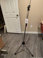 Full view of adjustable silver mic stand showing tripod base with rubber feet placed on floor, in room with wooden floor and beige walls.