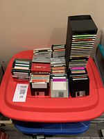 Photo showing a large assortment of 3.5-inch floppy disks stacked and spread over a red plastic container. Various colors and brands are visible, disks appear pre-owned.