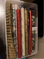 Multiple assorted Christmas-themed gift wrapping paper rolls arranged standing upright in a plastic container. Rolls display various holiday designs including stripes, floral, and holly patterns.