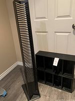 Photo of tall black metal vertical CD rack with multiple slots standing on floor next to closed white door.
