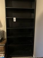 Front view of tall black bookshelf showing all 6 shelves empty, shelf pins visible for adjustability.