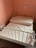 White metal IKEA child bed frame with vertical bars and swirl decoration on headboard and footboard with beige SULTAN SOVA mattress rolled on top.