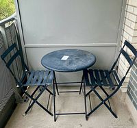 Full set of metal bistro table and two chairs on outdoor balcony. Table round and black with rust marks, chairs folding with metal slats.