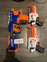Photo showing one blue Nerf Elite blaster with an orange circular drum magazine and two white Nerf Ultra blasters with orange and black detailing.