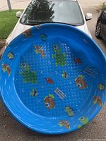 Photo of blue children's pool with cartoon fish and turtle decorations, placed on a driveway with a white car behind.
