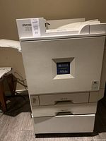 Full side view of the Ricoh Aficio CL7000 printer showing paper input and output trays, beige color and overall condition.