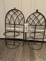 Two bronze metal wall planter holders with Gothic Arch design side by side, showing full structure including hanging loops and circular plant holder rings.