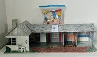 Full view of the Marx tin litho L-shaped ranch style dollhouse showing detailed interior and exterior artwork, chimney, and roofing with the bag of vintage furniture placed on top.