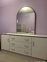 White dresser with three drawers and two side cabinets with metal handles, topped with an arched mirror, shown against a purple wall.