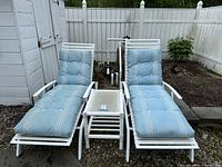 Two white metal chaise lounges with blue cushions and a white metal framed glass top side table between them, set outdoors on gravel next to a white fence.