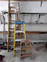 Three ladders standing next to each other against a wall in a workshop or garage space. The 6-foot tall wooden stepladder is behind the yellow fiberglass 4-foot stepladder and the small 2-foot wooden stepladder is in front.