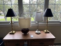 Four vintage lamps shown on a wooden table in front of a window, including two brass base lamps with black shades, one ceramic base lamp with beige shade, and one metal base lamp with white pleated shade.