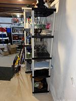 Side view of the shelving unit showing four black plastic shelves supported by white legs, with various glass items on shelves (not included).