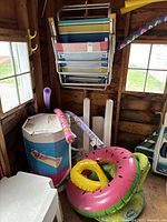 Wide angle photo showing several stacked foldable beach chairs, inflatable pool floats in pink and yellow, a box with inflatable lounge bed, and foam pool noodles in a wooden shed.