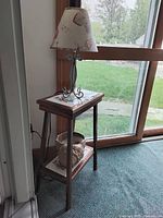 Side view of wood side table with ceramic tile tops holding a lamp with floral shade and ceramic seashell on bottom shelf, placed near window.