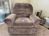 Front view of the brown fabric recliner rocker chair in a living room corner with framed pictures around.