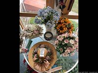 Photo of multiple floral arrangements, a floral decorative hat, and a candle on a glass table.