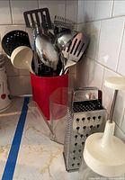 Photo showing stainless steel kitchen tools including slotted spoons, serving spoons, potato masher, slap chopper, gravy separator, and a metal box grater arranged on a countertop with a red utensil holder.