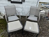Photo showing two gray mesh patio chairs with metal frames and armrests placed side by side outdoors on gravel next to a building, each paired with a footstool with a light gray cushion.