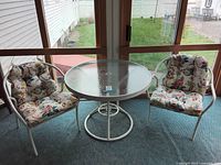 Photo of a round glass outdoor table with two white metal chairs with floral cushions in a sunroom or enclosed porch setting.