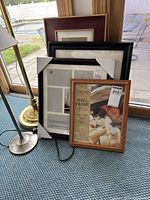Four assorted picture frames stacked and displayed on a carpeted floor near a window and two lamps, showing wood and black finishes.