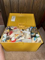 Open yellow plastic sewing box showing spools of thread, needles, Velcro, and other sewing supplies inside