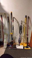 Photo showing eight cleaning tools arranged side by side including mops, brooms, and steam cleaners against a white wall on a carpeted floor.