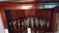 Photo of wooden cabinet shelf with a collection of vintage cordial glasses and other drinking glasses arranged in rows.