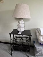 Wrought iron table with ceramic lamp on top, shown against wall and carpeted floor.