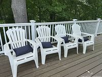 Four white Adirondack chairs lined up on a wooden deck railing with black cushions on each seat.