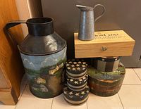Front view of all items showing the tole art milk can with painted houses, small cream pitcher on wooden cheese crate, wine box on Americana nesting boxes