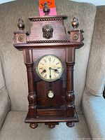 Front view of the wooden antique wall clock showing decorative top and carved face sculpture, pendulum and Roman numeral dial.