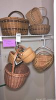 Seven handcrafted baskets of various shapes, sizes, and weave patterns arranged on metal shelving and hanging on a rod. Different textures and colors of woven wicker.