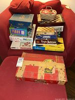 Lot of vintage board games and an Erector set box on red chair, showing boxes including Monopoly, Taboo, Yahtzee, Jeopardy, Triple Yahtzee, and others with wear visible.