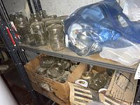 Metal shelving holding multiple clear glass canning jars and cardboard boxes with other jars and supplies. Items appear dusty and in used condition.
