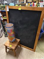 Large framed chalkboard with a cylindrical map poster sitting on a small wooden stool in a storage setting.