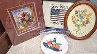 Four pieces of embroidery and needlepoint art, including floral designs and a patriotic flag motif, framed and loose in a bag.