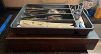 Farberware cutlery knives with wooden handles and other utensils in a plastic cutlery tray on a wooden surface