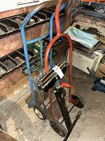 Two metal hand trucks, one blue and one red, placed together in a basement workshop area with shelving and various tools around. The red hand truck is in front, alongside a black lumber roller stand with two wheels and a handle.