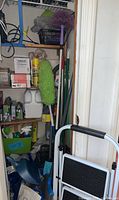 Shelves and floor corner with cleaning tools, green microfiber duster, mop handles, plastic step stool, broom and dustpan.