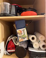 Shelf containing assorted kitchen appliances including Dash waffle maker, a red handheld mixer, boxed apple corer, vacuum sealer rolls and other small appliances.