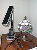 Two lamps on a wood table: a stained glass style lamp with floral shades and an OttLite desk lamp with black base and arm.