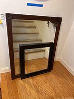 Two vintage wood framed wall mirrors, one large with carved corner details and one smaller with simple black frame, placed against a wall and floor