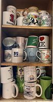 Shelf filled with assorted mugs including souvenir, humorous, and novelty designs